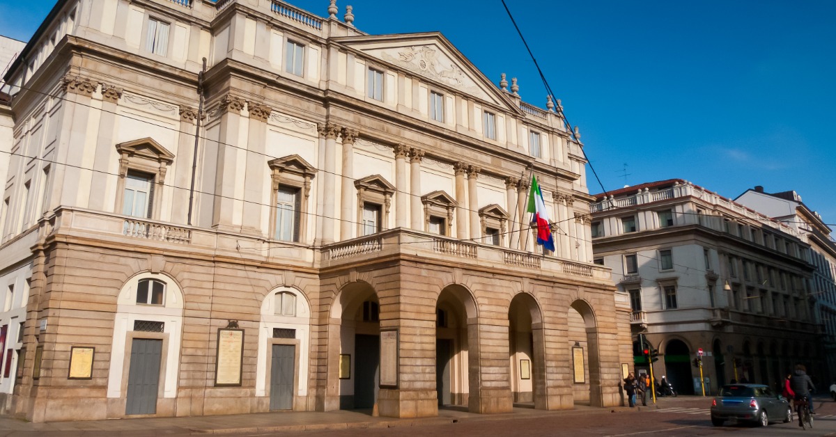 The Teatro alla Scala in Milan, a building with ornate columns, arched entrances, and flags displayed under a clear blue sky.